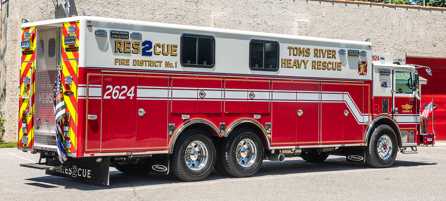 Officer’s side of a Pierce Walk-In Heavy Duty Rescue Fire Truck parked outside on a sunny day next to a building. 