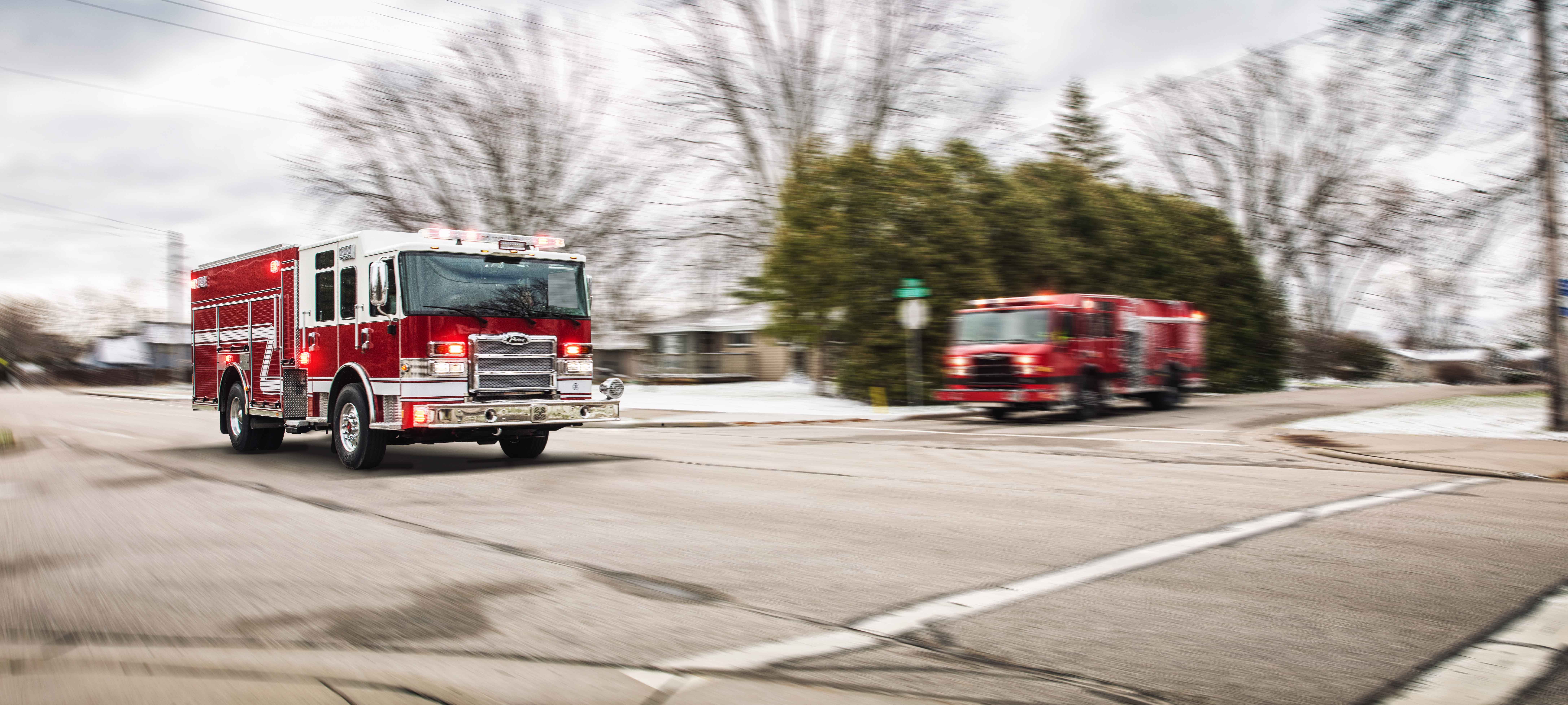 Two Pierce Fire Trucks driving in the street at an intersection on a cloudy day. 