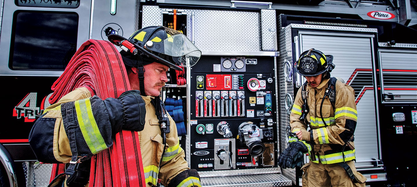 Two firefighters pulling hose and equipment off their Pierce aerial ladder truck