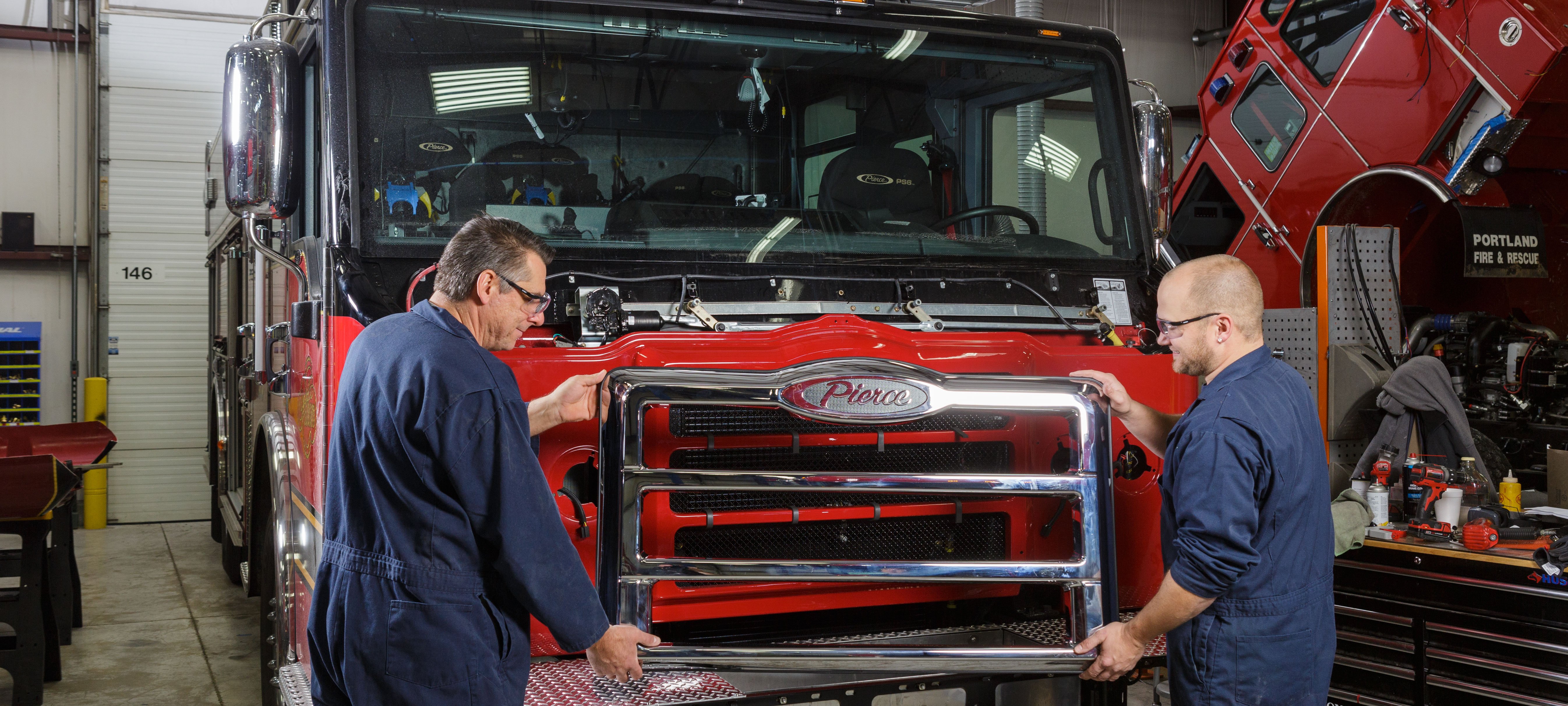Two individuals putting parts on the front of a Pierce Fire Truck parked inside of a warehouse.  