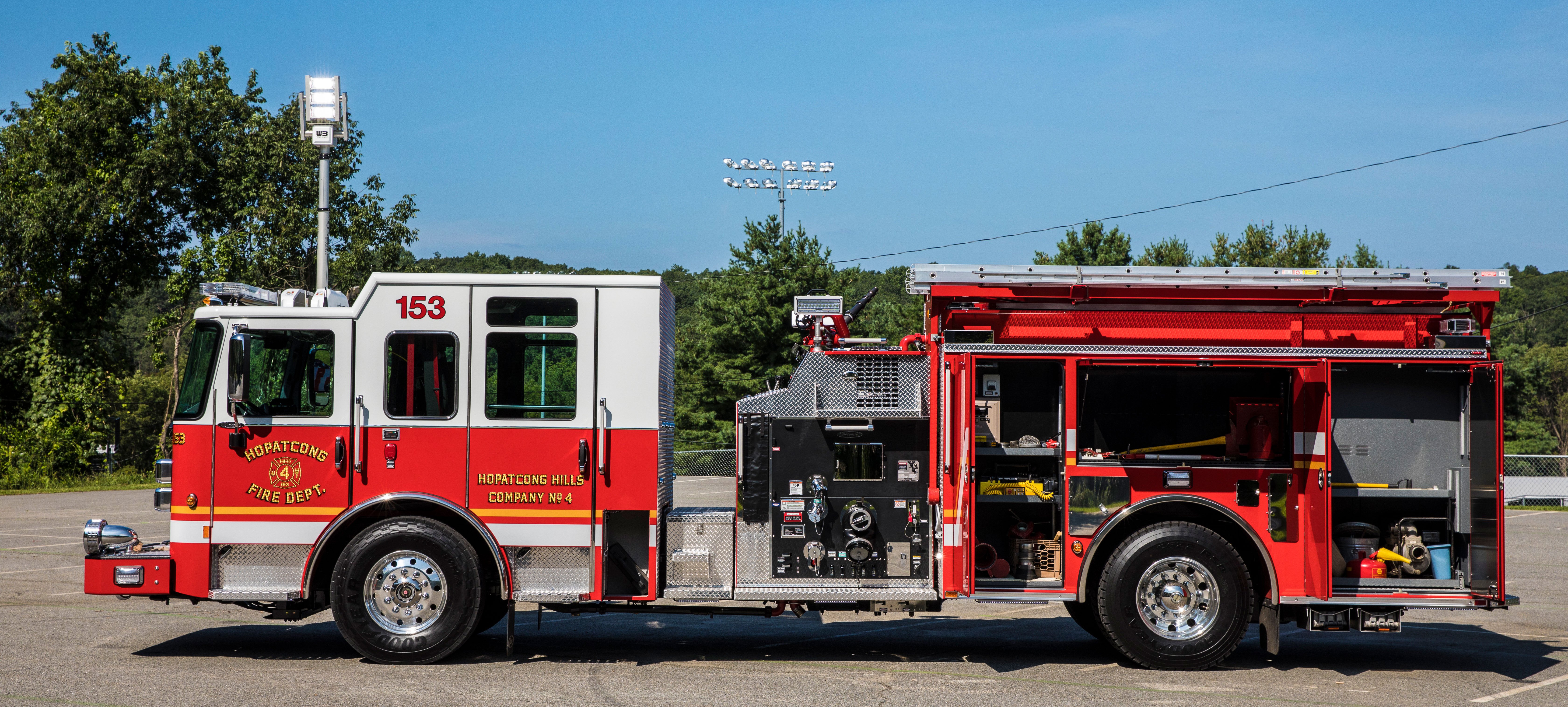 Driver’s side of a Pierce Pumper Fire Truck parked outside in a parking lot with side compartments open showing storage space.  