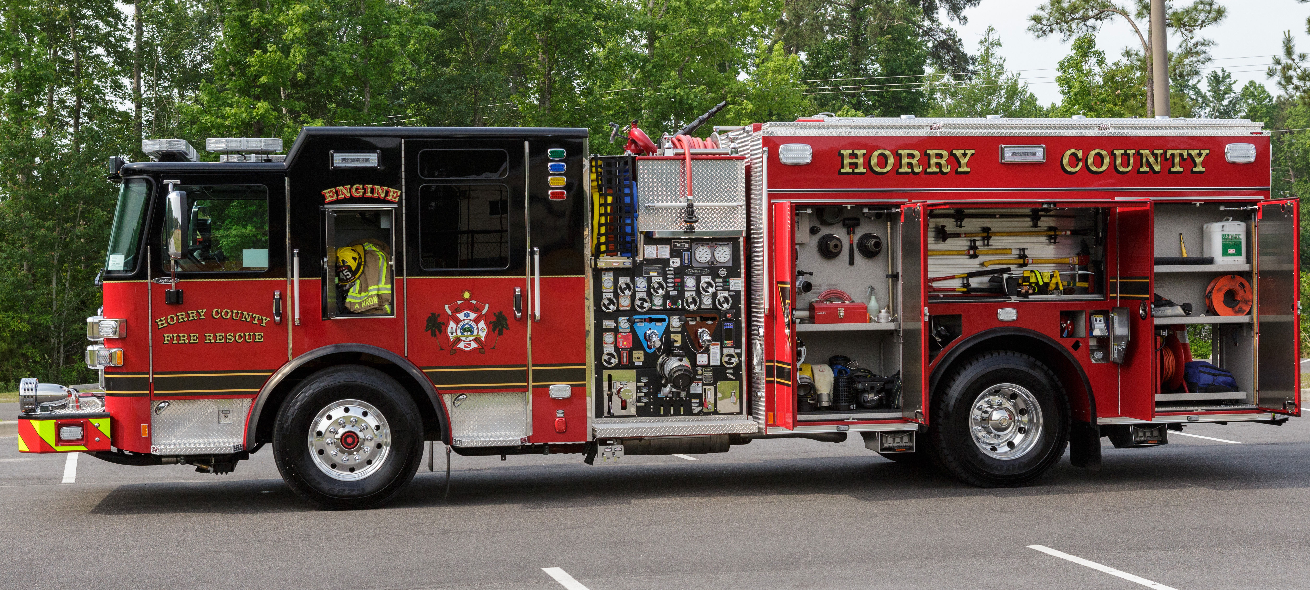 Officer’s side of a Pierce Pumper Fire Truck parked in a parking lot with compartments open showing storage space.  