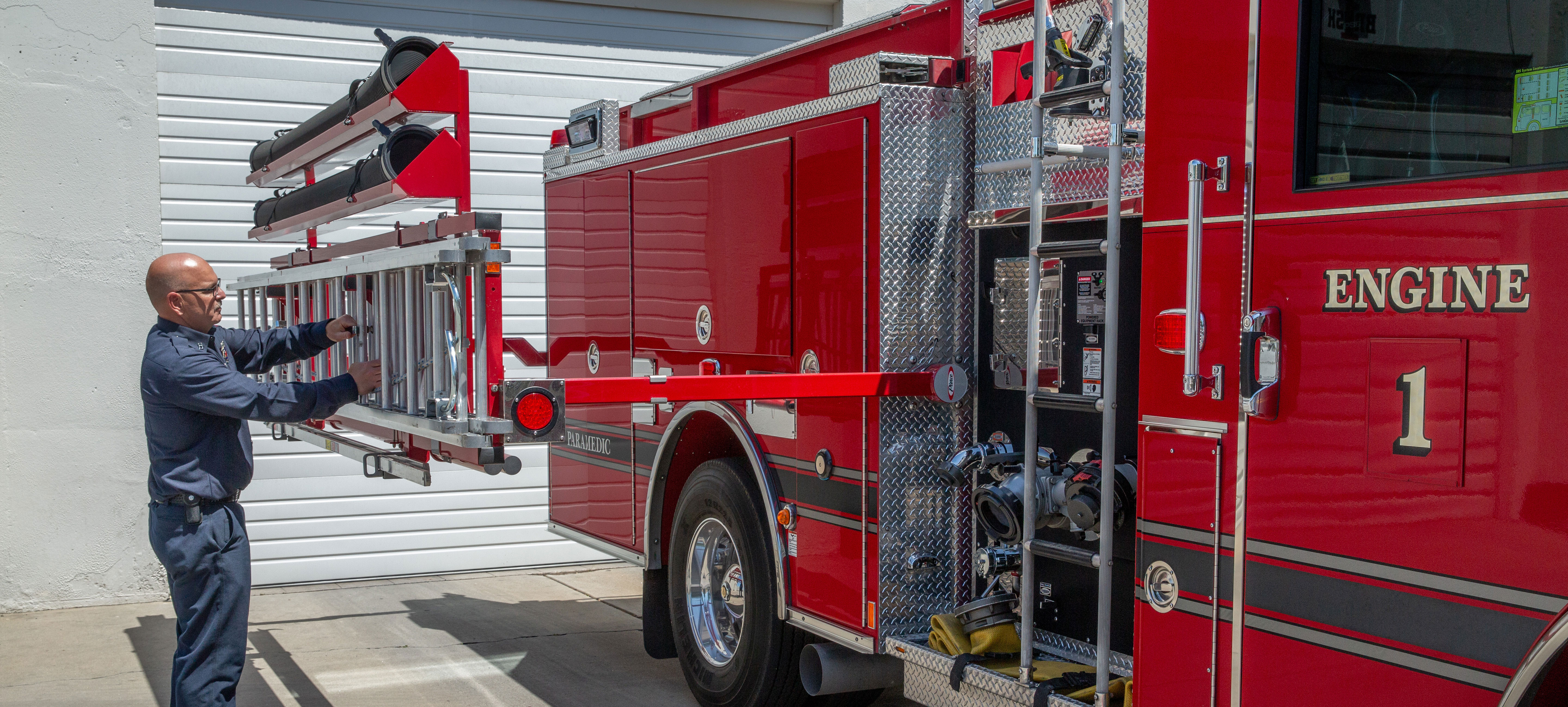 A man putting a ladder on the passenger’s side of a Pierce Pumper Fire Truck Ladder rack parked outside on a sunny day. 