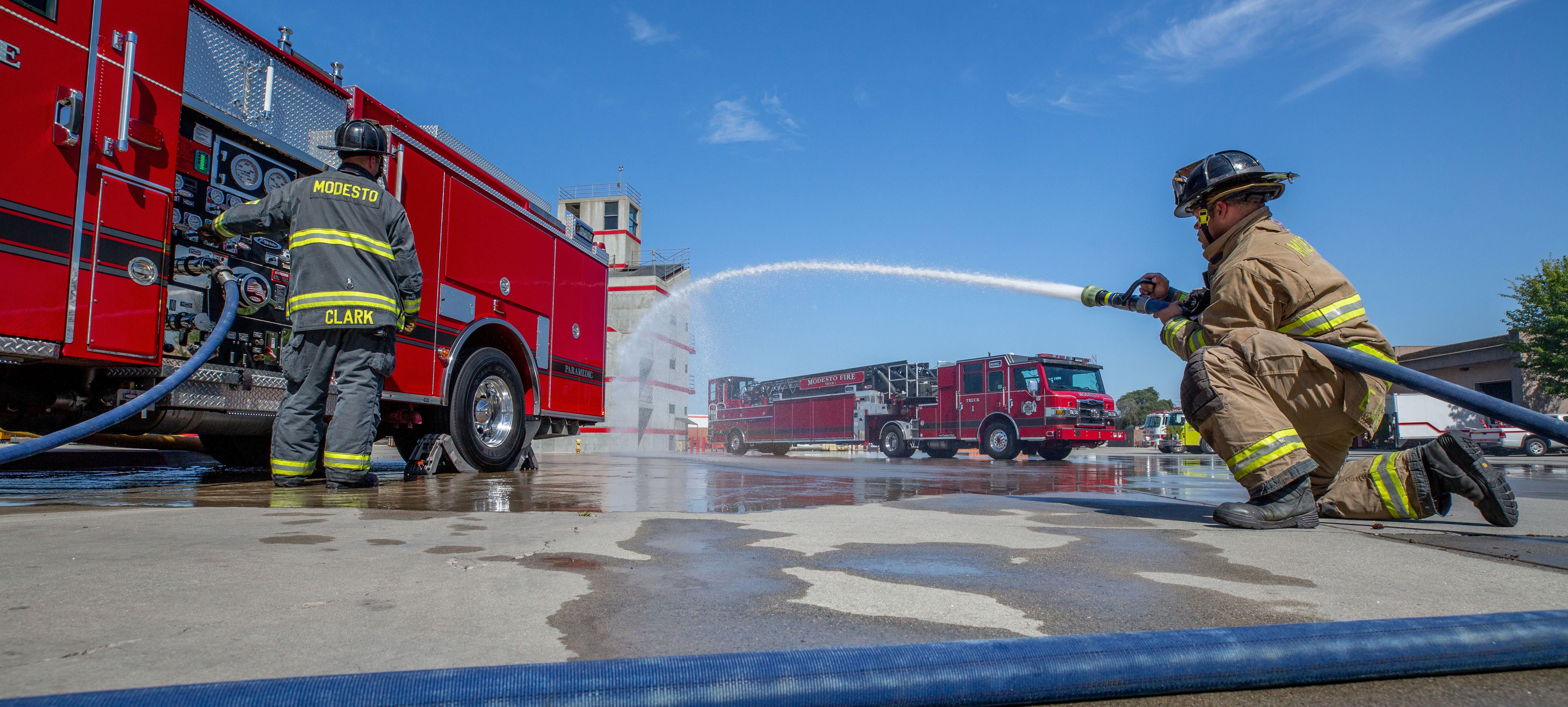 A firefighter standing at the pump panel and a firefighter operating a hose spraying water from a Pierce Pumper Fire Truck parked outside.  