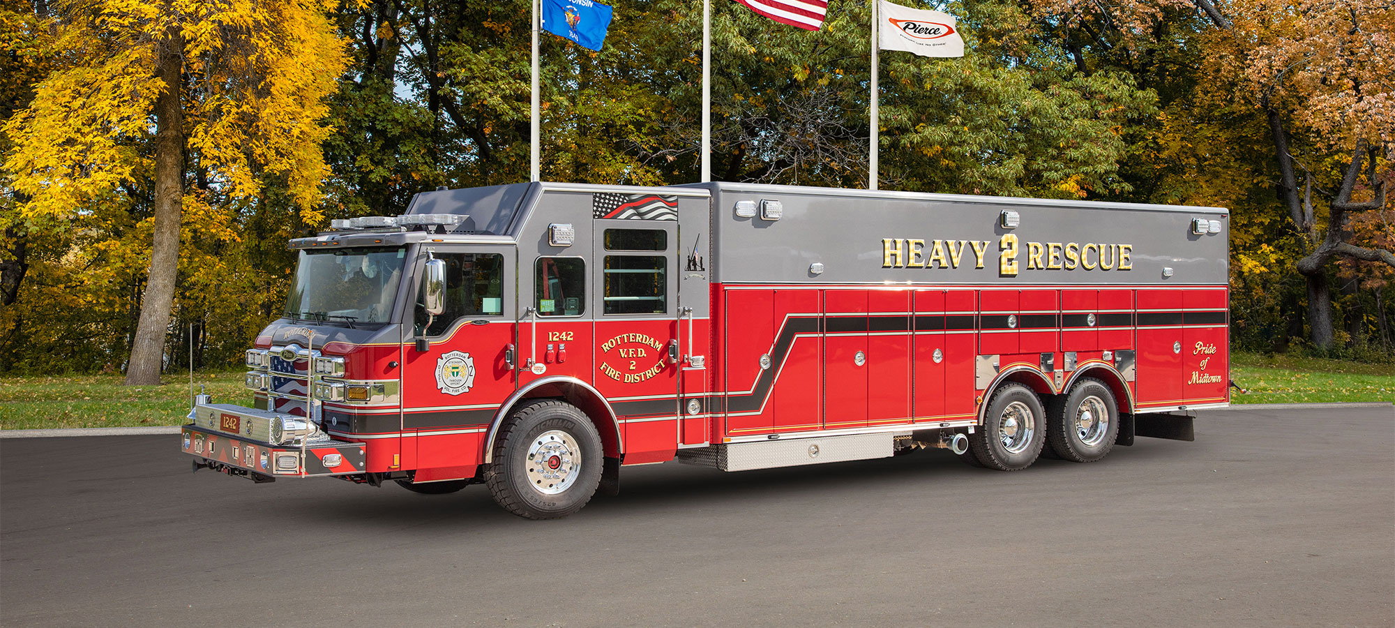 A Pierce Heavy-Duty Walk-In Rescue fire truck parked in front of flag poles on a fall day.