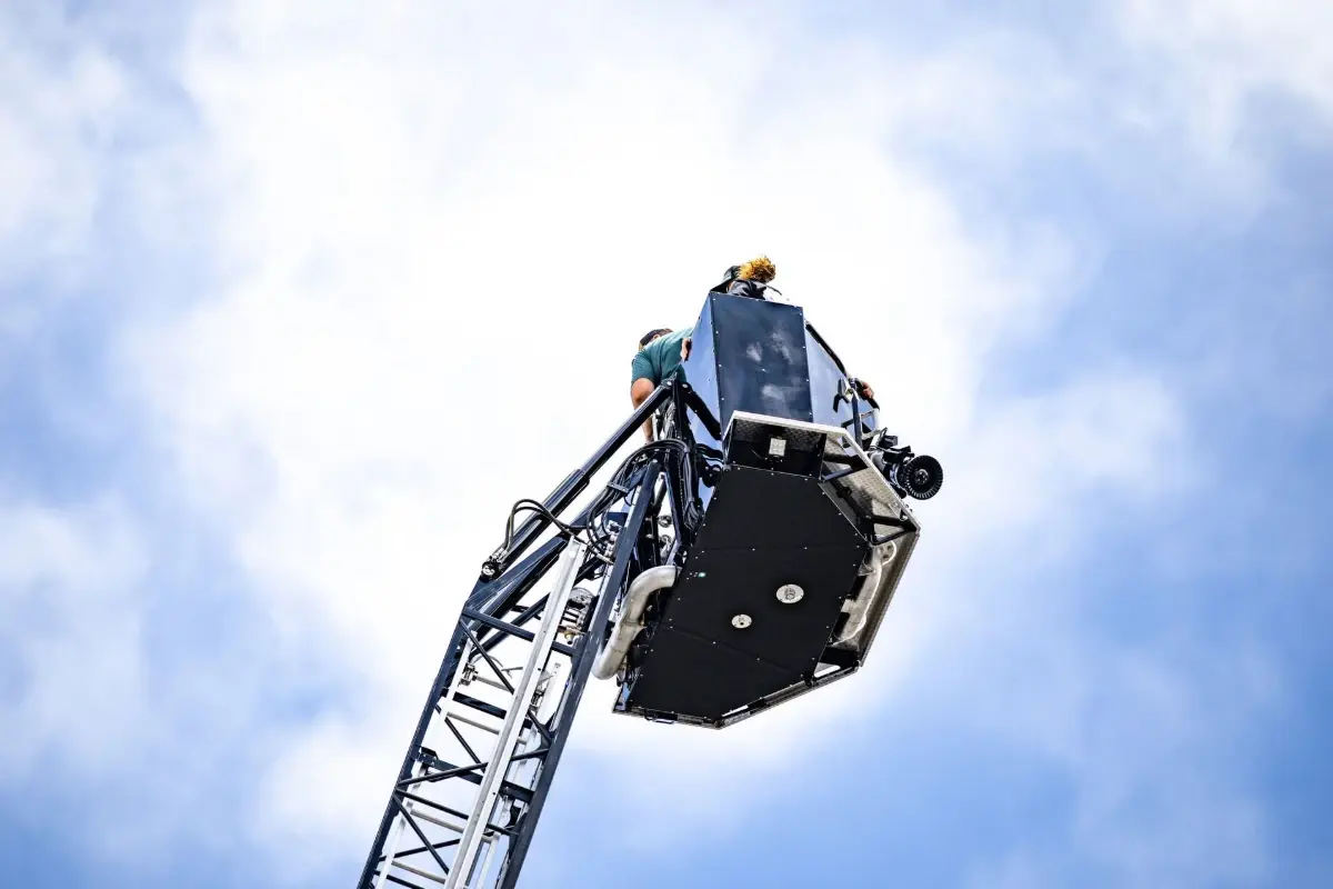 Firefighters stand in a Pierce fire truck aerial platform basket against a cloudy blue sky.