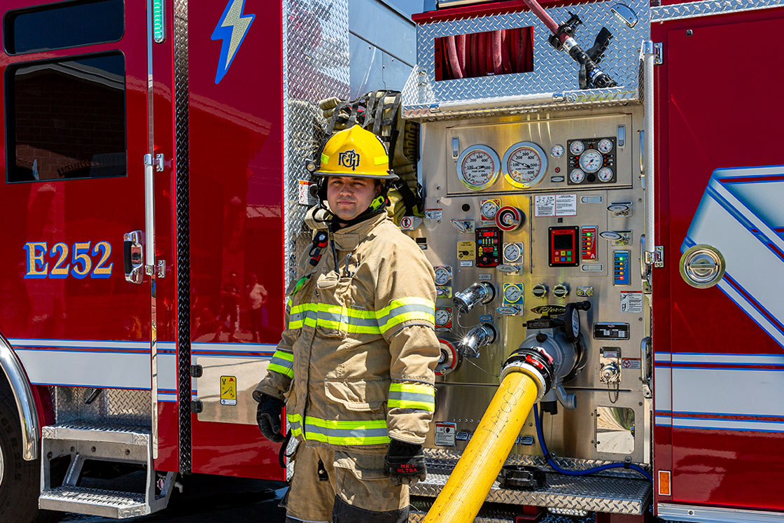 A firefighter in turnout gear standing beside a pump panel with a charged hose coming from the pump.