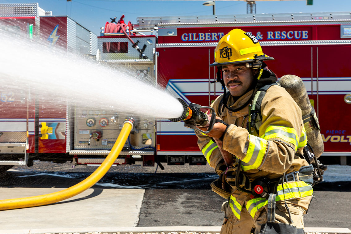 A firefighter in turnout gear standing in front of a fire truck spraying water from a fire hose.