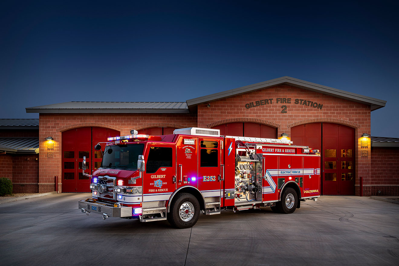The front drivers side of a Velocity Volterra Pumper with the lights on in front of a fire station at night.