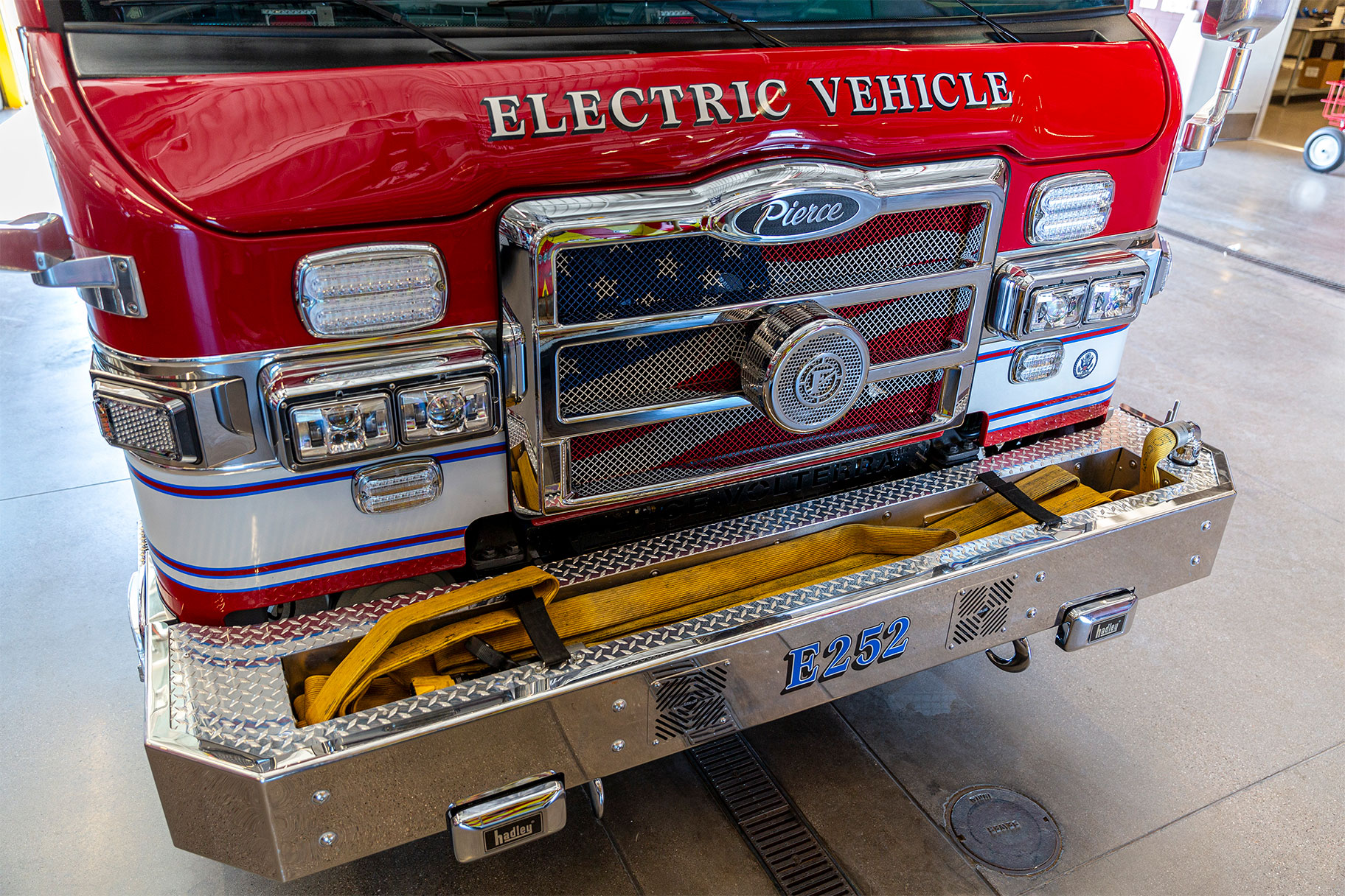 The front of a Volterra electric fire truck parked in a firehouse with hose in the bumper.