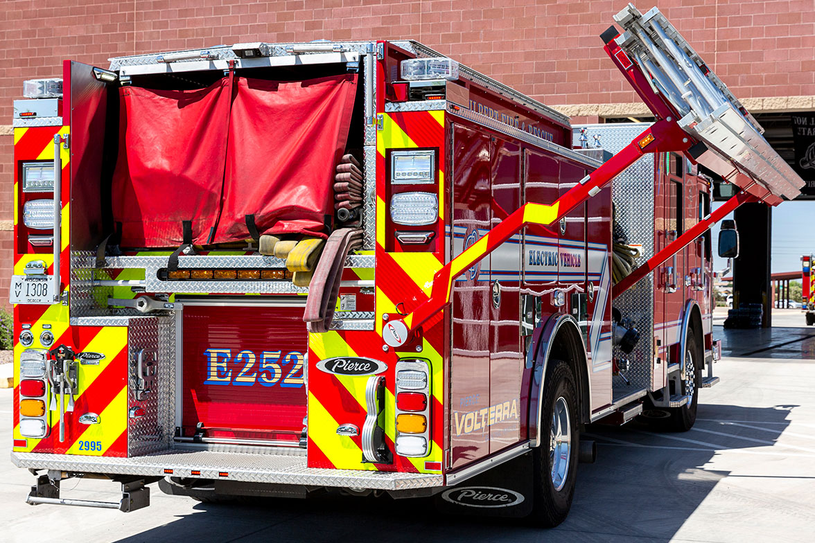 The officer's side rear of a fire truck with the hydraulic ladder rack coming down with ground ladders outside of a fire station.