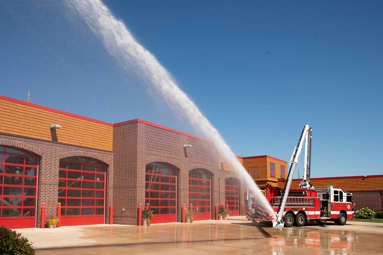 Pierce Fire Truck parked outside on a sunny day in front of a Fire Station with the High-Reach Snozzle extended out in front of the truck spraying water into the air. 