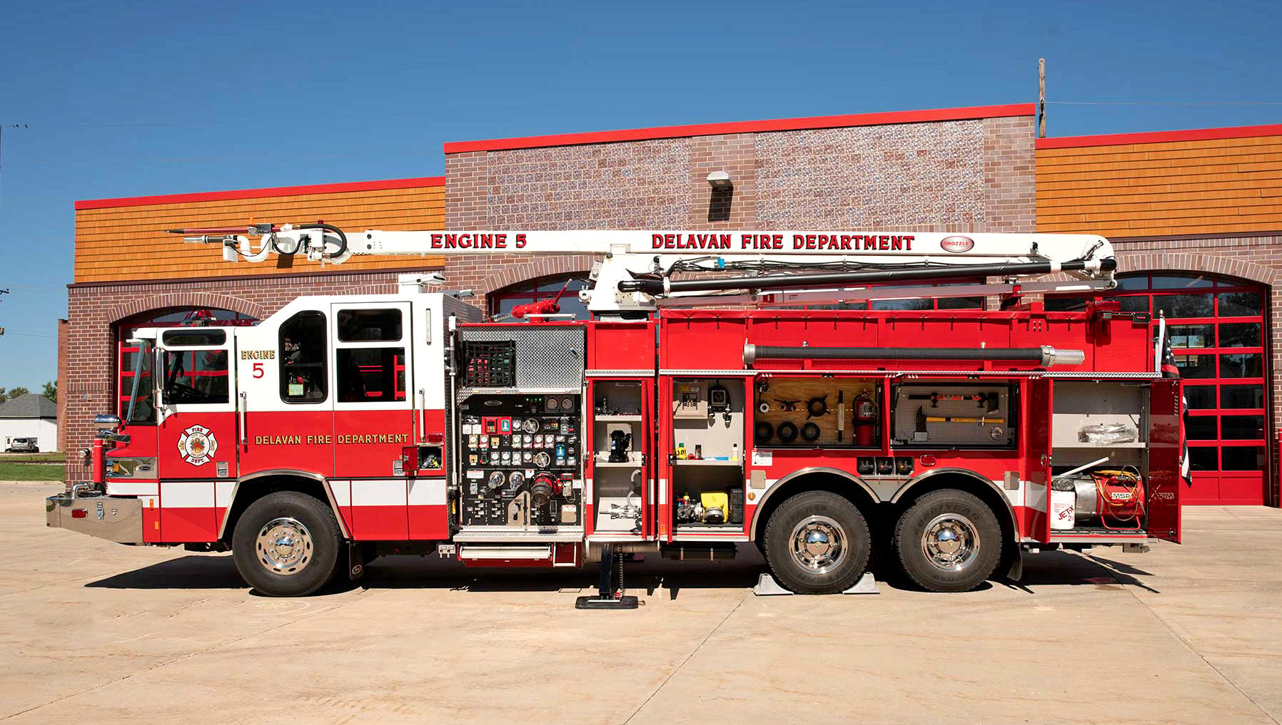 Driver’s side of a Pierce Fire Truck with a high-reach extendable snozzle parked outside on a sunny day in front of a Fire Station with compartment doors open showing storage space. 