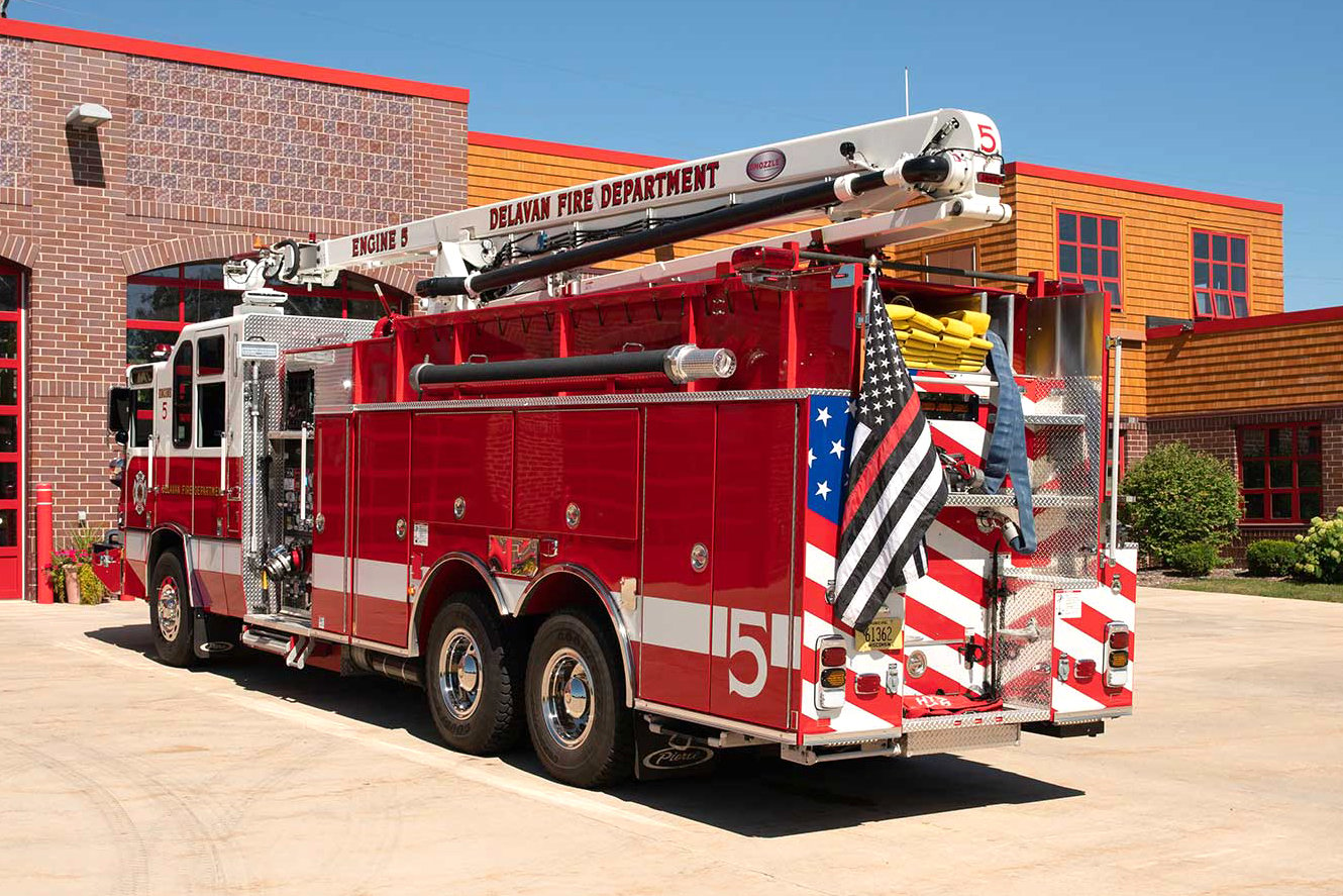 Driver’s side and rear of a Pierce Fire Truck with a snozzle parked in front of Fire Station on a sunny day. 