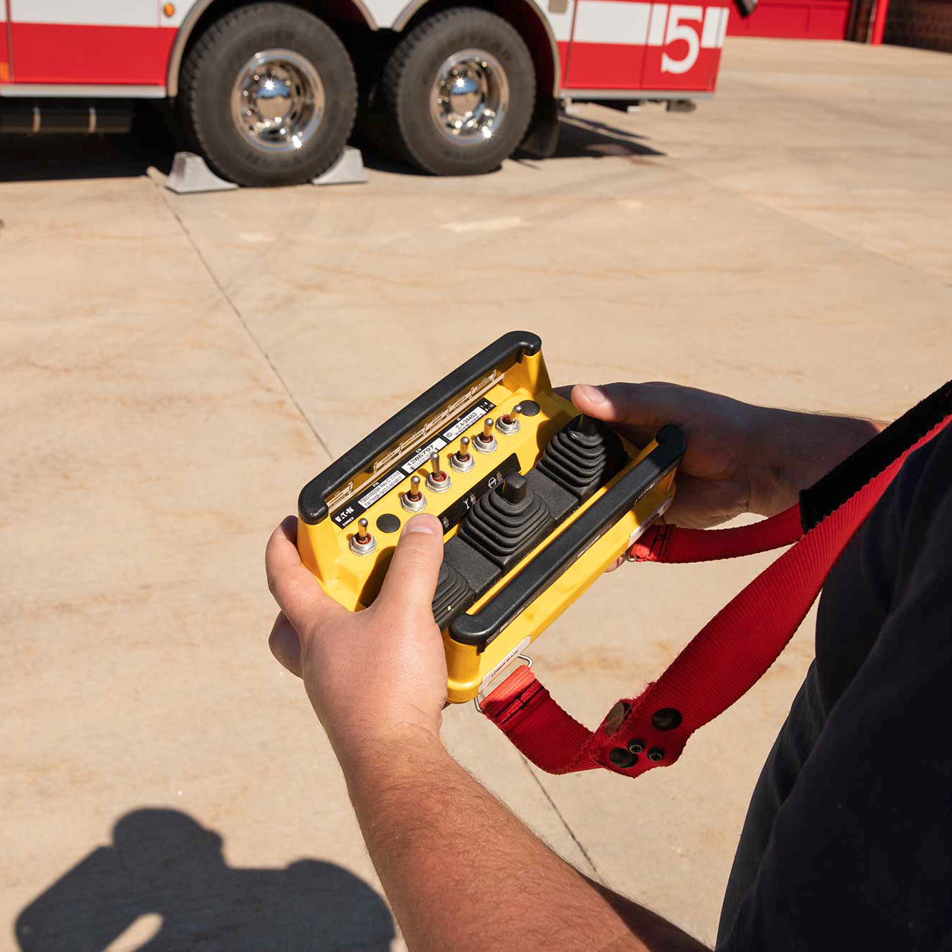 A man standing near a Pierce Fire Truck parked outside on a sunny day holding a remote control for a Fire Truck Snozzle. 