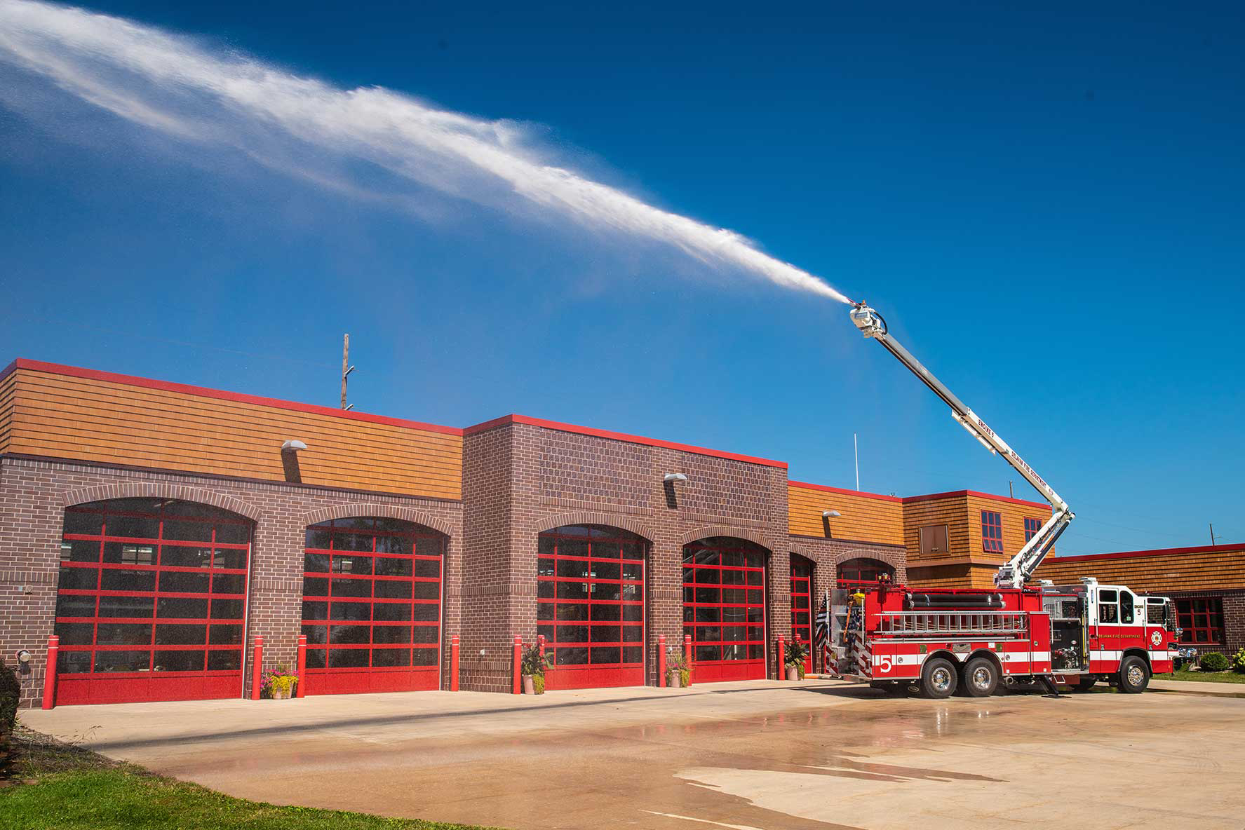 A Pierce Fire Truck parked outside on a sunny day in front of a Fire Station with its High-Reach Snozzle extended into the air spraying water. 