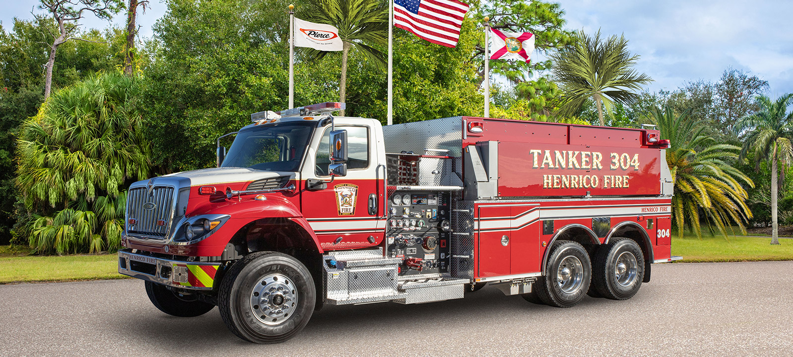 Front and Driver’s side of a Pierce Commercial Dry Side Tanker for Henrico Fire parked outside near trees and three flags on a sunny day. 