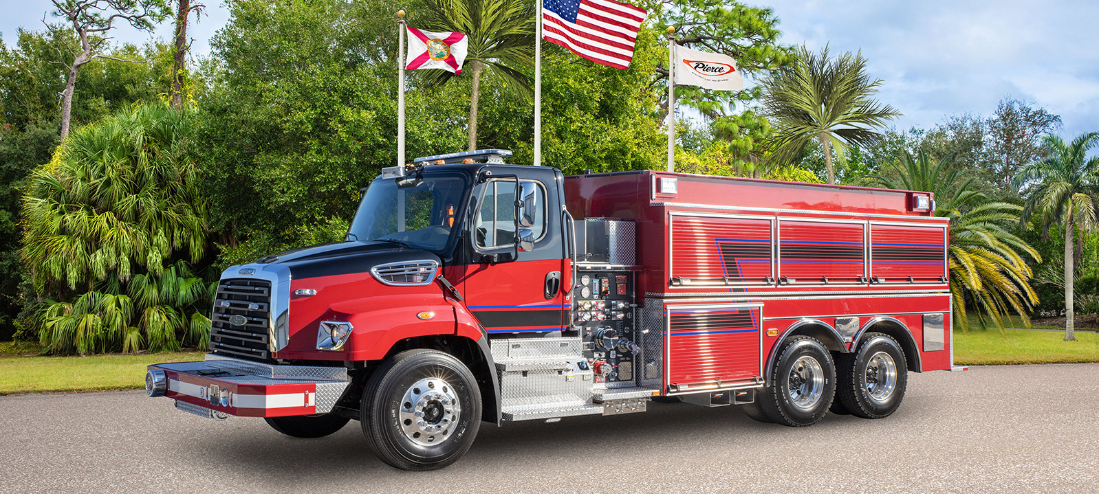 Front and driver’s side of a Pierce Commercial Wet Side Tanker Fire Truck parked outside near trees and three flags on a sunny day. 