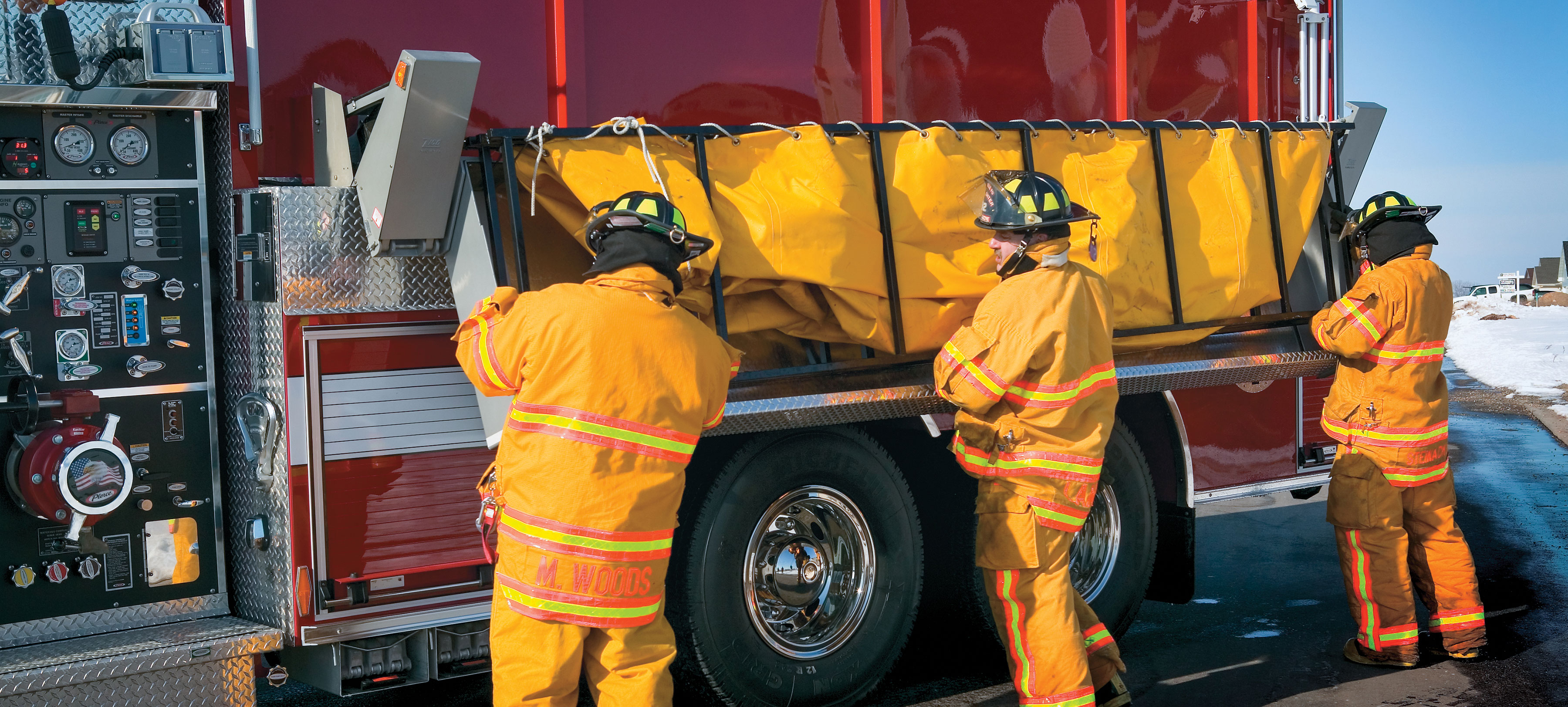 Three firefighters working on a Pierce Tanker Fire Truck parked outside in a parking lot with swing-down folding storage on the driver’s side. 