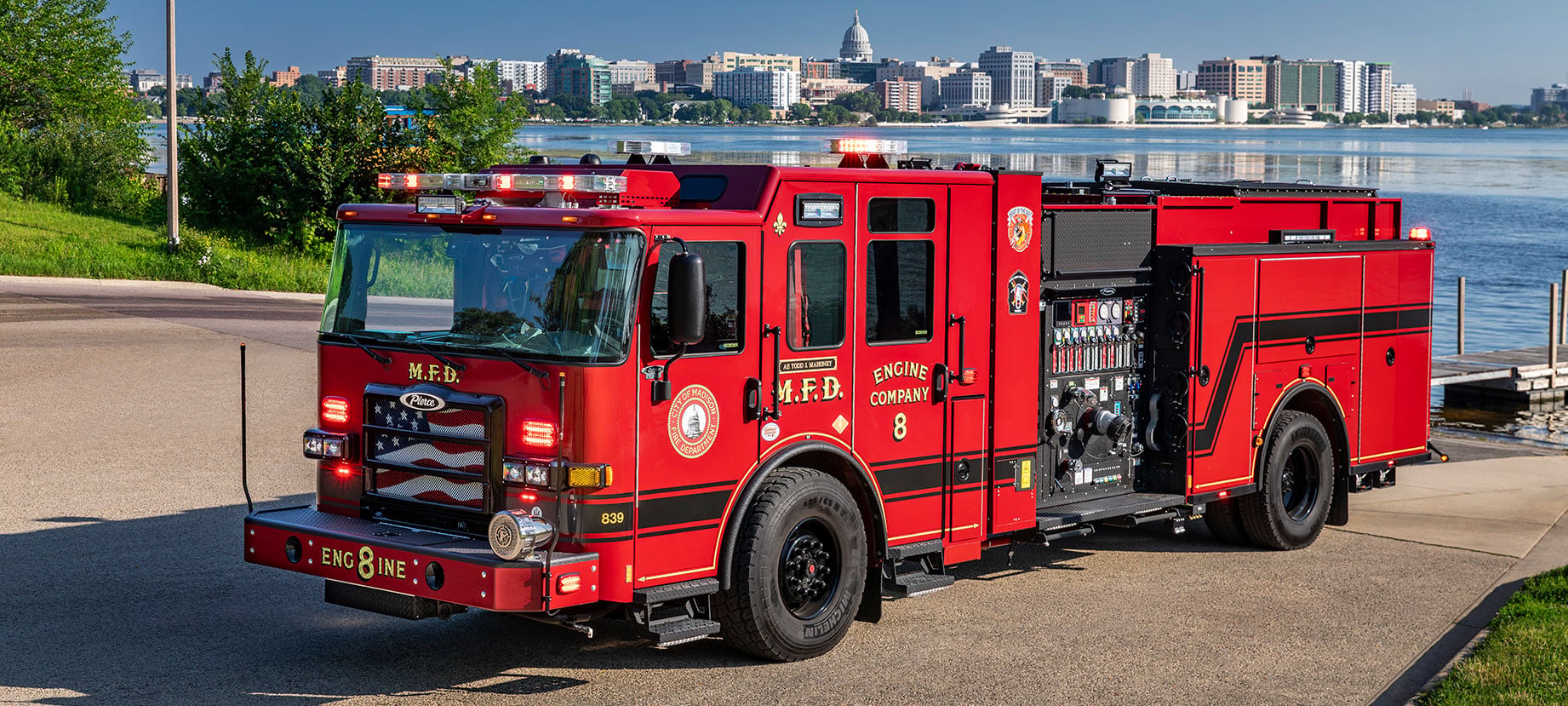 The front driver's side of a Pierce Volterra Electric Fire Truck with the emergency lights on parked in front of a lake with a city in the background.