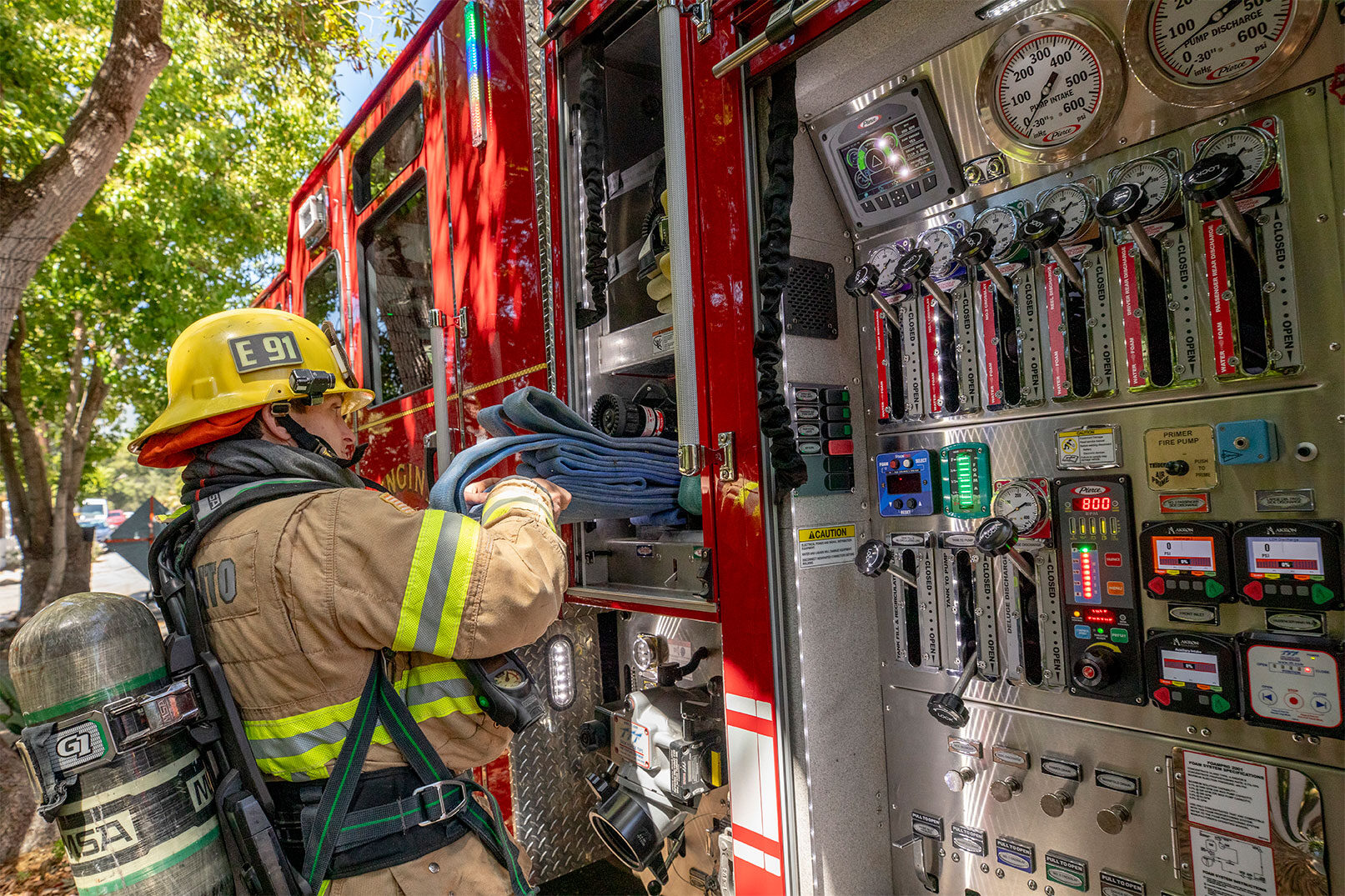 A firefighter in turnout gear pulling fire hose out of a crosslay on a fire truck next to a pump panel.