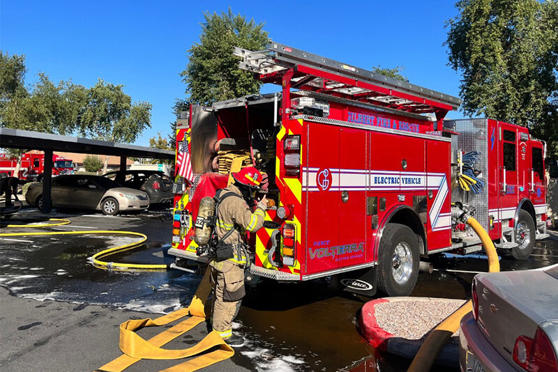 Red electric pumper fire truck with a white stripe is parked while firefighters deploy hose at an emergency scene.