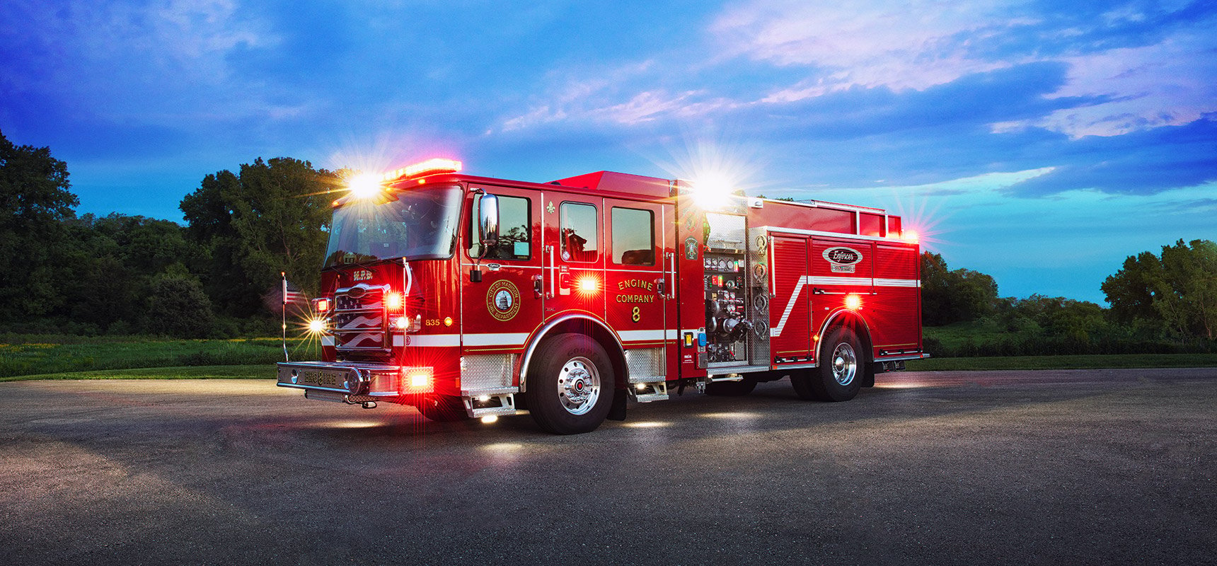 A red electric fire truck has its warning lights on at dusk with a blue evening sky in the background. 